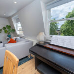 Modern loft conversion with bright living space, cozy sofa, piano, and large windows overlooking greenery.